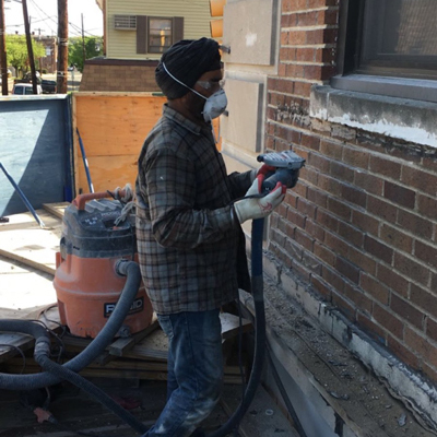 A man wearing a mask and gloves operates a vacuum cleaner to remove dust from a brick wall's surface.